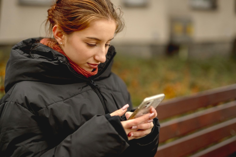 A woman in a black winter coat and red scarf stands outdoors near a bench, focused on her smartphone. The image illustrates how group texting services can deliver timely event and meeting reminders, even on the go.