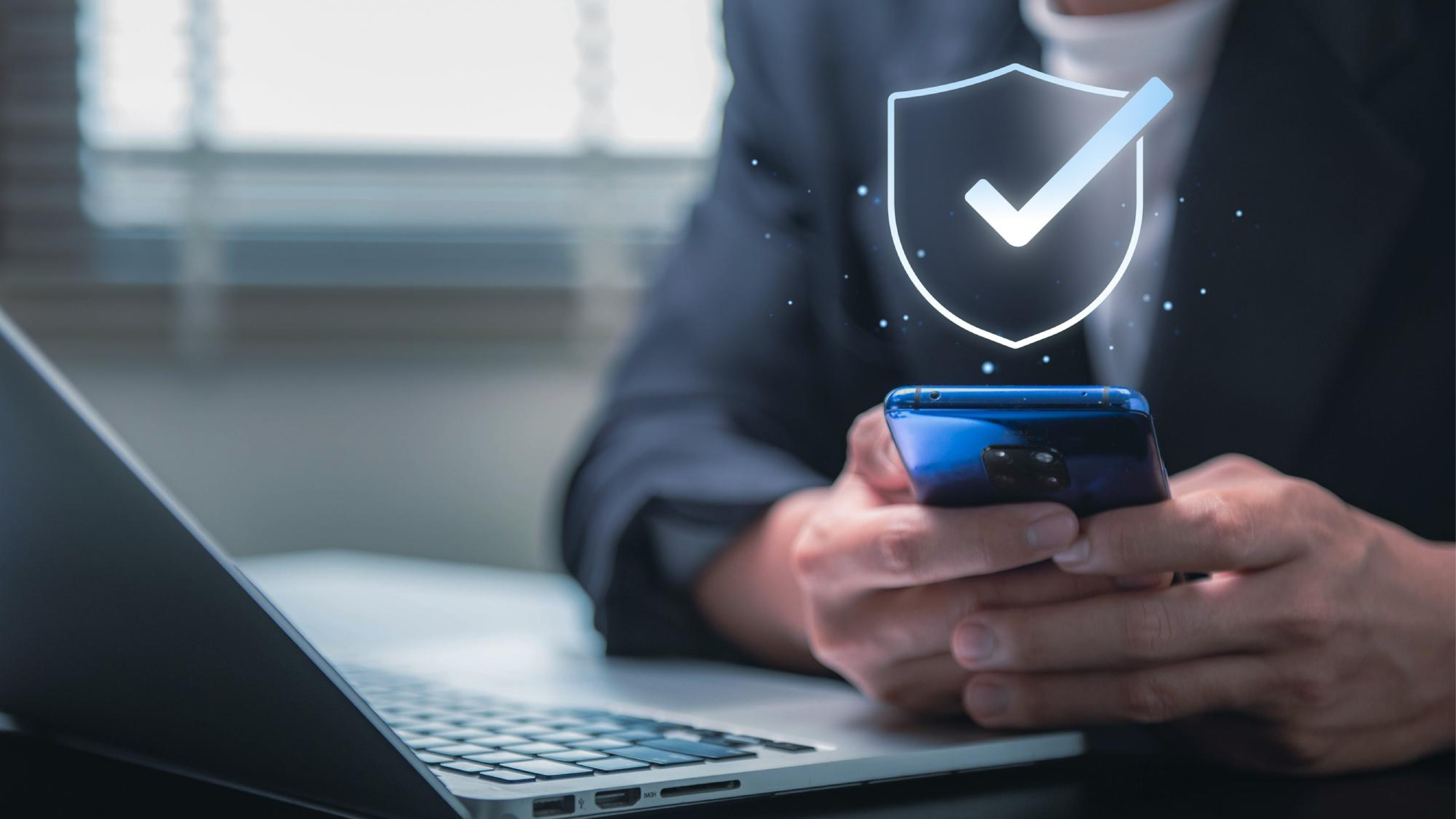 Close-up of a person holding a smartphone in front of a laptop, with a digital shield and checkmark icon overlayed, symbolizing secure SMS marketing software designed to prevent spam text messages.
