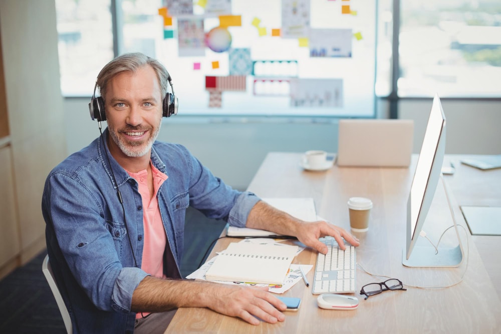 A smiling man with gray hair and headphones sits at a computer desk in a bright office. His relaxed posture reflects the ease of managing remote work using group texting services for fast team communication.