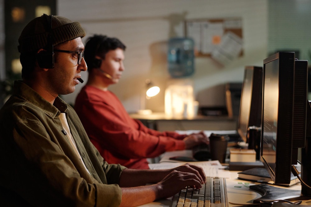 Two customer support agents wearing headsets work at desktop computers in a dimly lit office. The scene highlights the reliability of group texting services to support 24/7 communication in high-demand environments.