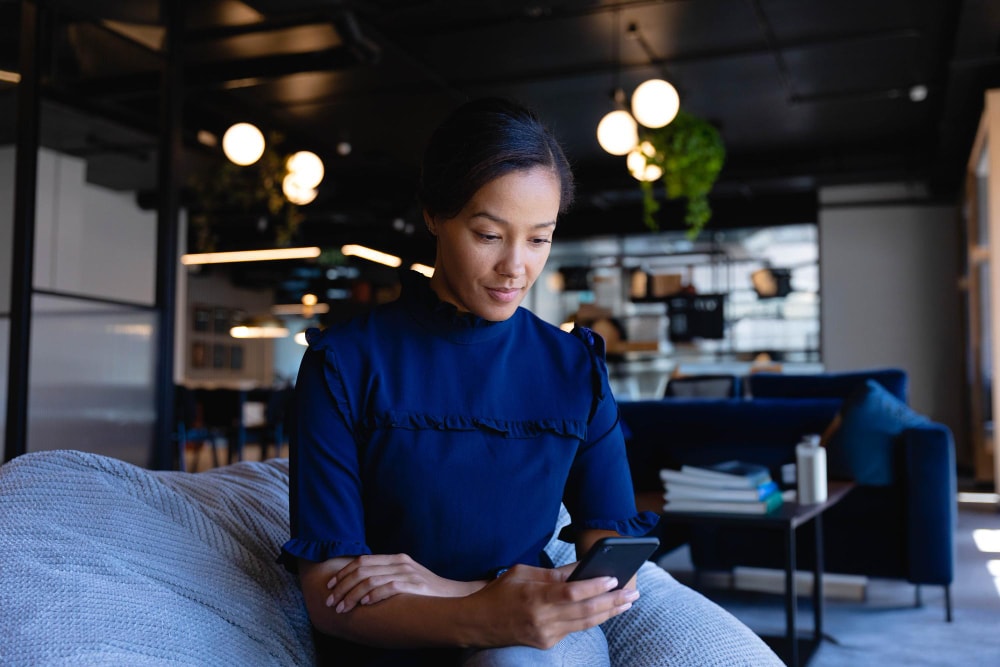 A woman in a navy blouse sits in a modern office lounge, looking at her smartphone. The stylish, relaxed environment reflects how group texting services can support professional communication from anywhere.