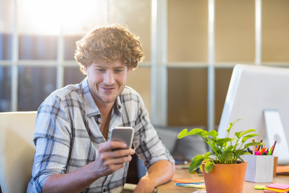 A man in a plaid shirt smiles while reading a message on his smartphone at his desk, where a potted plant and office supplies are visible. The image captures a casual moment of using group texting services for quick updates.