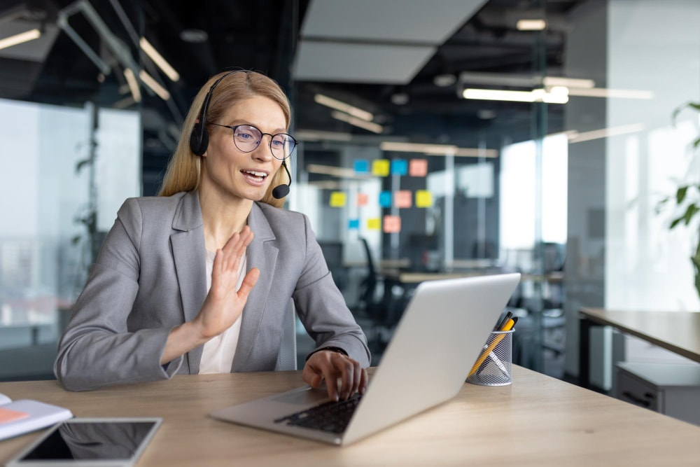 A professional woman wearing glasses and a headset smiles and waves during a video call on her laptop in a modern glass-walled office. She uses group texting services to enhance virtual collaboration and engagement.