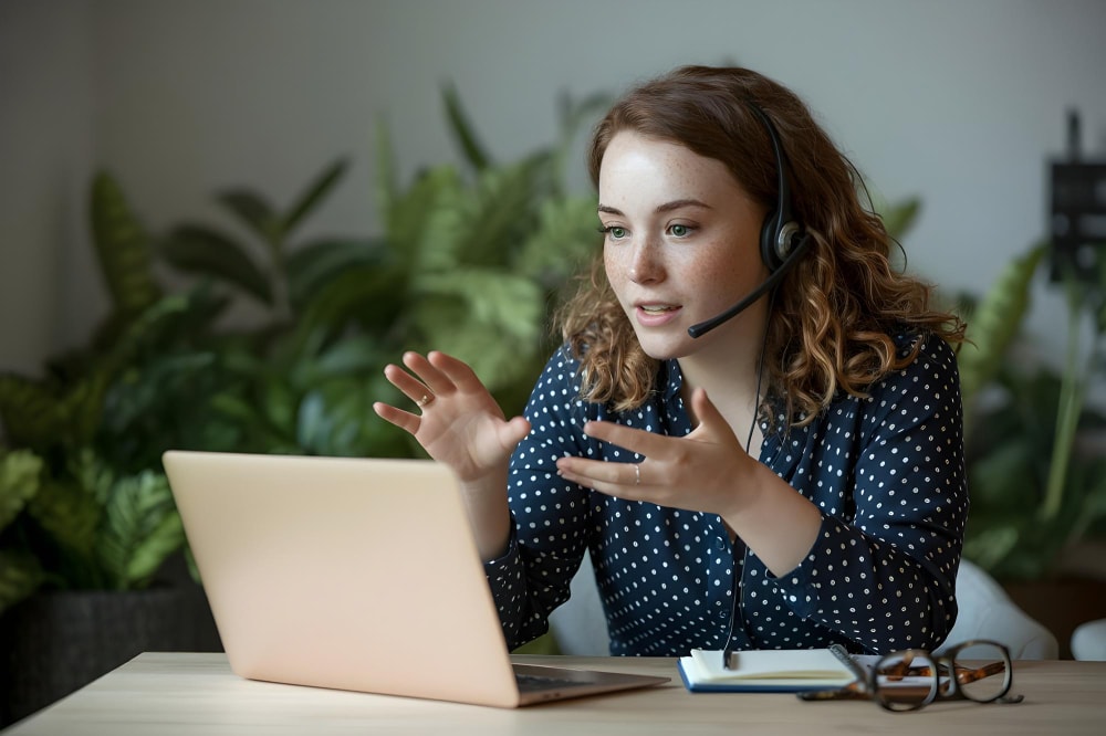A woman with curly red hair and a headset gestures during a video call on her laptop, surrounded by greenery in a home office. She is actively using group texting services to maintain real-time communication with her team.