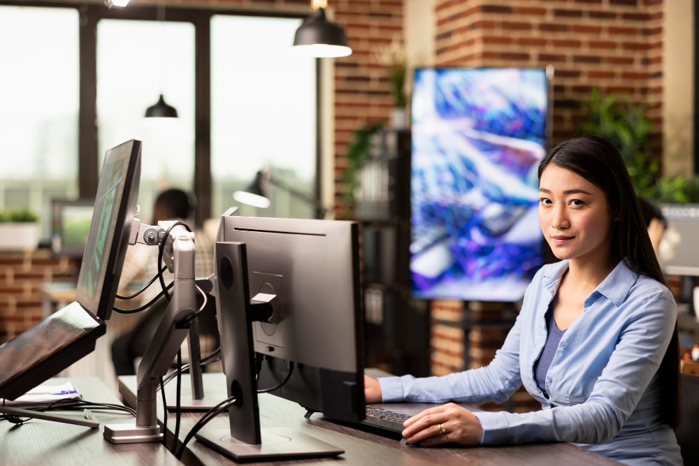 A woman in a blue shirt sits at a workstation with multiple monitors in a tech-style office. Her focused expression suggests she's managing internal communication through group texting services for streamlined operations.