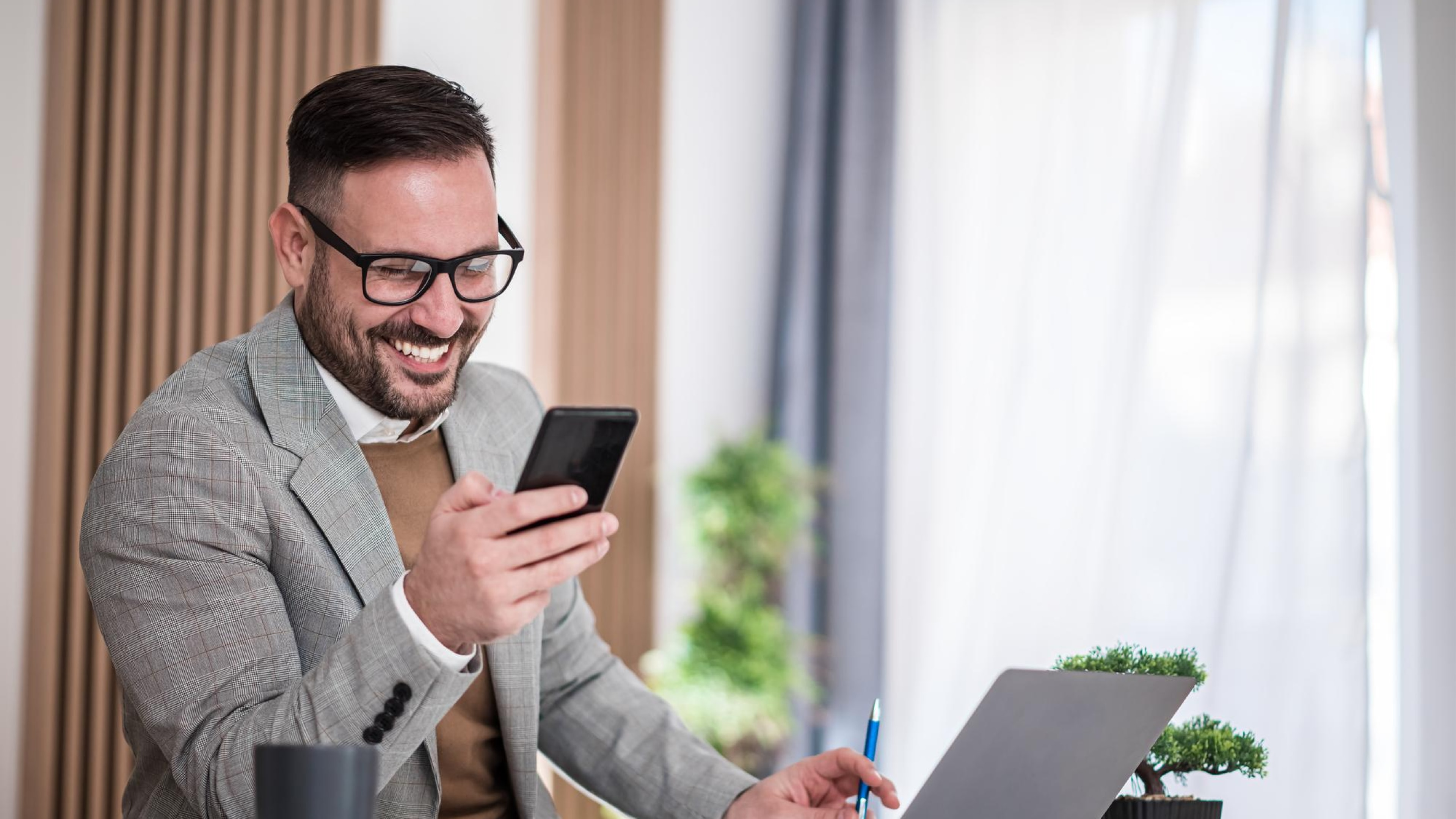 Businessman smiling at his phone while seated at a desk with a laptop, representing SMS marketing platforms that include clear opt out instructions for recipients.